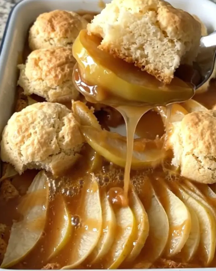 A warm, golden brown pear cobbler in an enameled baker with a serving spoon lifting a portion.