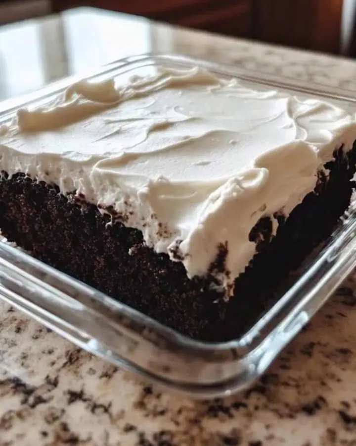 Square chocolate wacky cake with tall swirled white frosting in a clear glass dish on a granite countertop