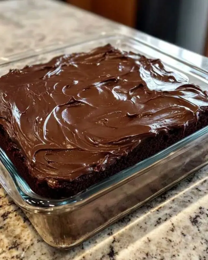 Gâteau au chocolat carré glacé d’un fondant brillant, dans un moule en verre posé sur un plan de travail en granit