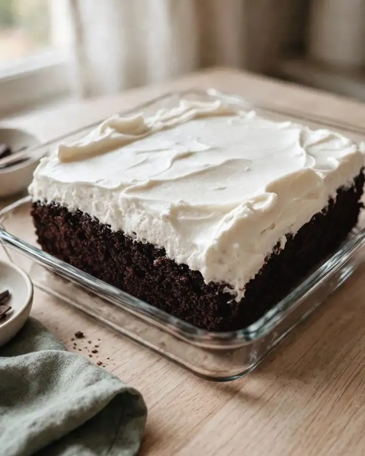 Gâteau au chocolat noir ultra-moelleux surmonté de glaçage blanc onctueux dans un plat carré en verre sur granit.