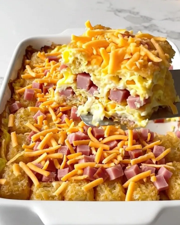 A top-down view of a rectangular ceramic baking dish filled with potato tots, ham, eggs, and melted cheddar cheese.