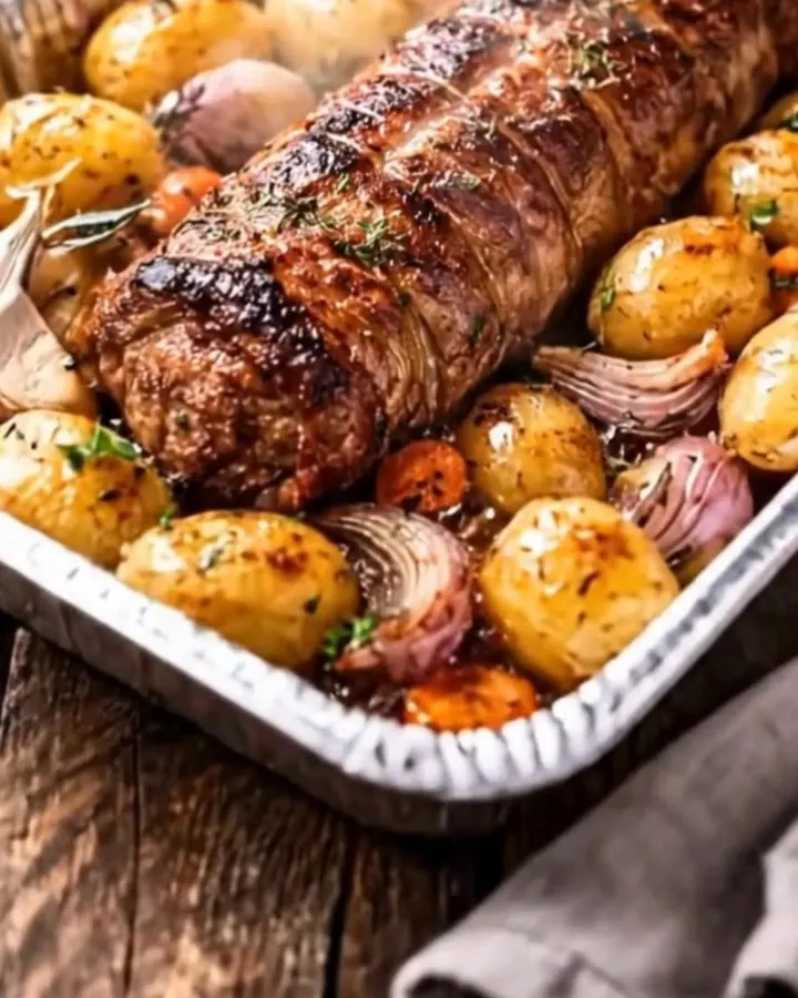 Rôti de porc doré reposant sur des grenailles dorées et des oignons rouges caramélisés dans un plat à rôtir, fumant