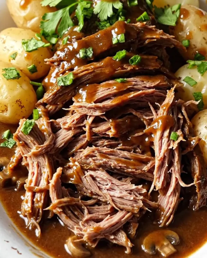 A close-up of a slow-cooked beef chuck roast in a gravy with potatoes, garnished with fresh green parsley.