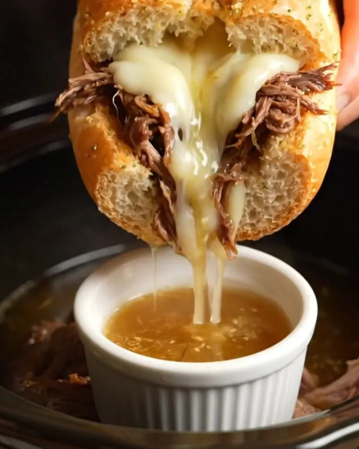 A close-up shot of a French Dip sandwich being dipped into a small ramekin of rich, dark au jus.