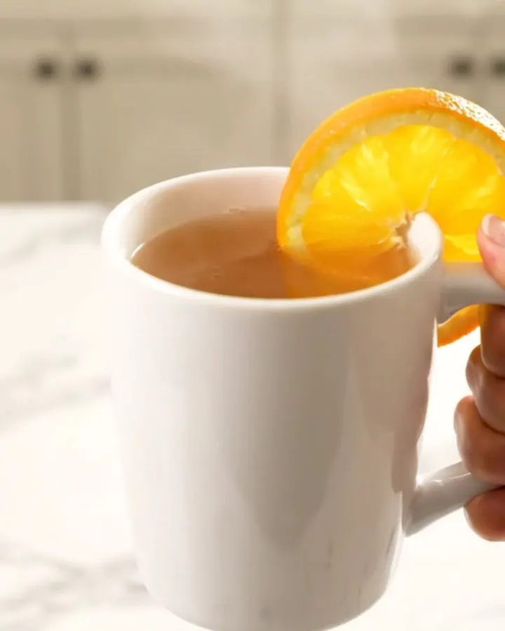 A white ceramic mug filled with steaming hot orange cinnamon wassail, garnished with an orange slice.