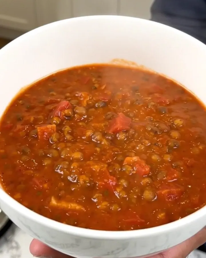 A top-down view of a black slow cooker filled with raw red lentils, diced carrots, onions, and tomato coulis.