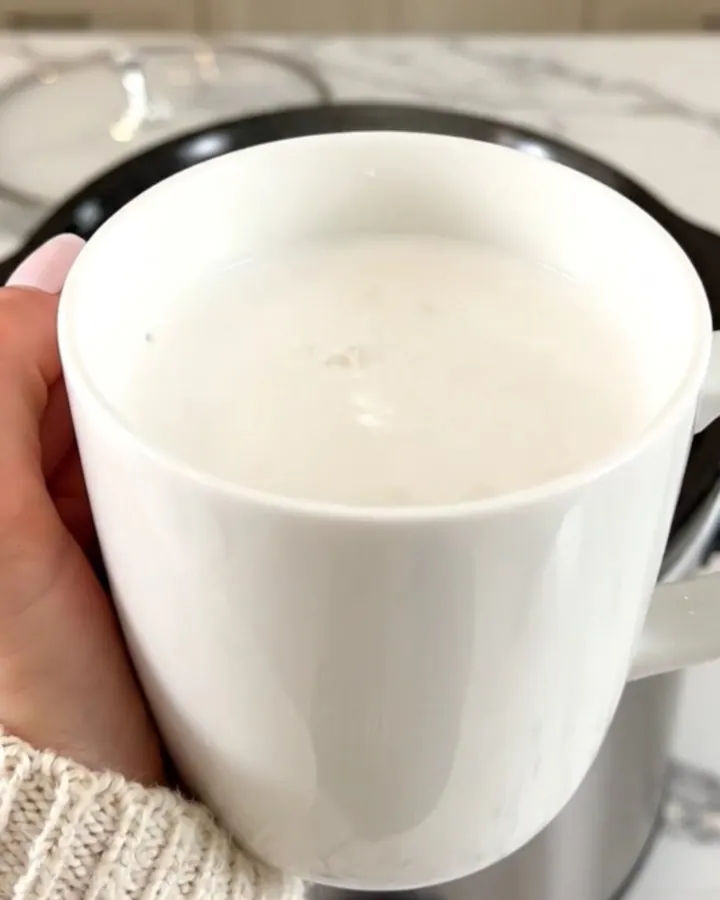 A white ceramic mug filled with creamy white hot chocolate on a white speckled countertop.