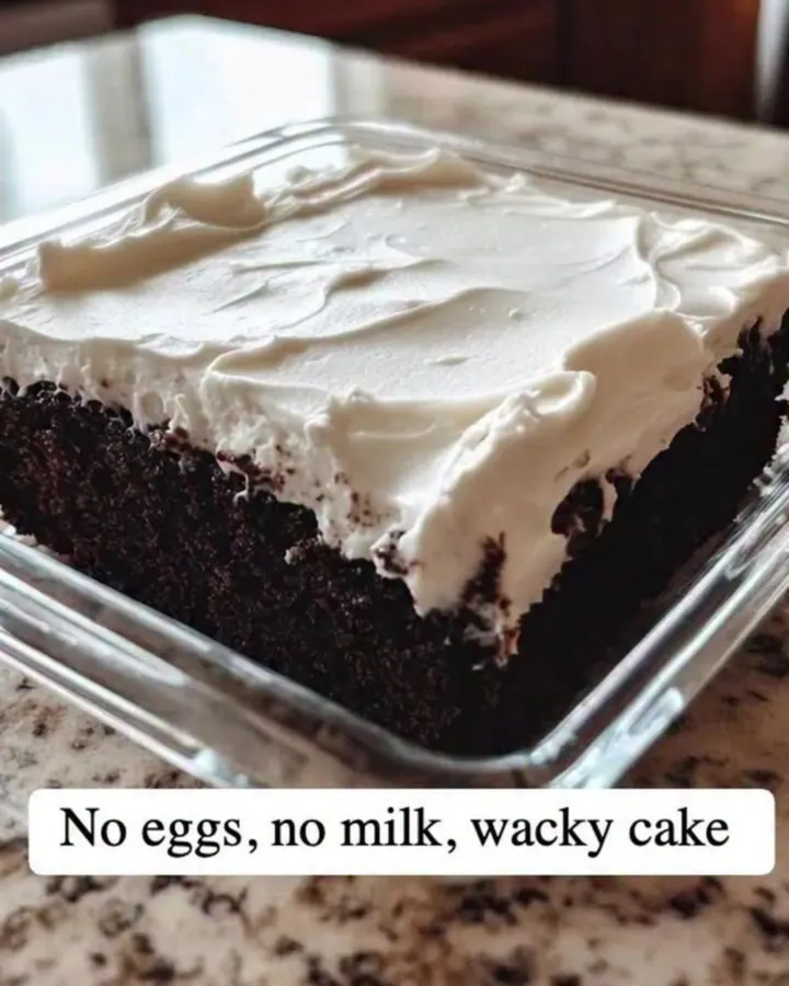 Fork cutting through white vanilla frosting into a moist dark chocolate wacky cake in a square glass dish